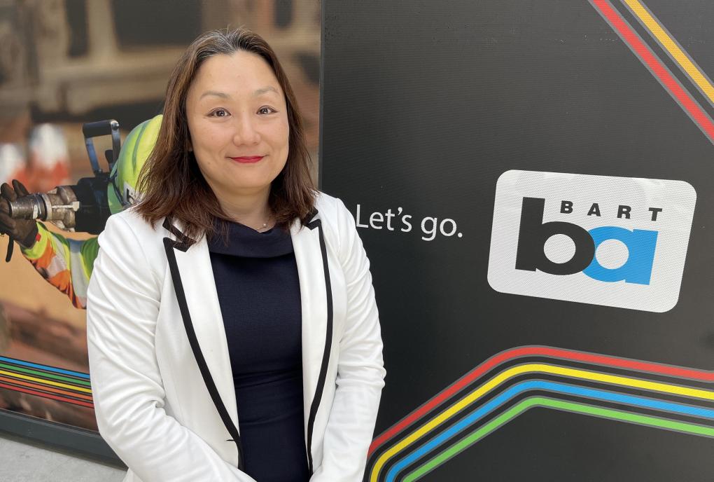 Ni Lee in a black shirt and white blazer pictured in front of a slice of the BART map with the blue and black BART logo