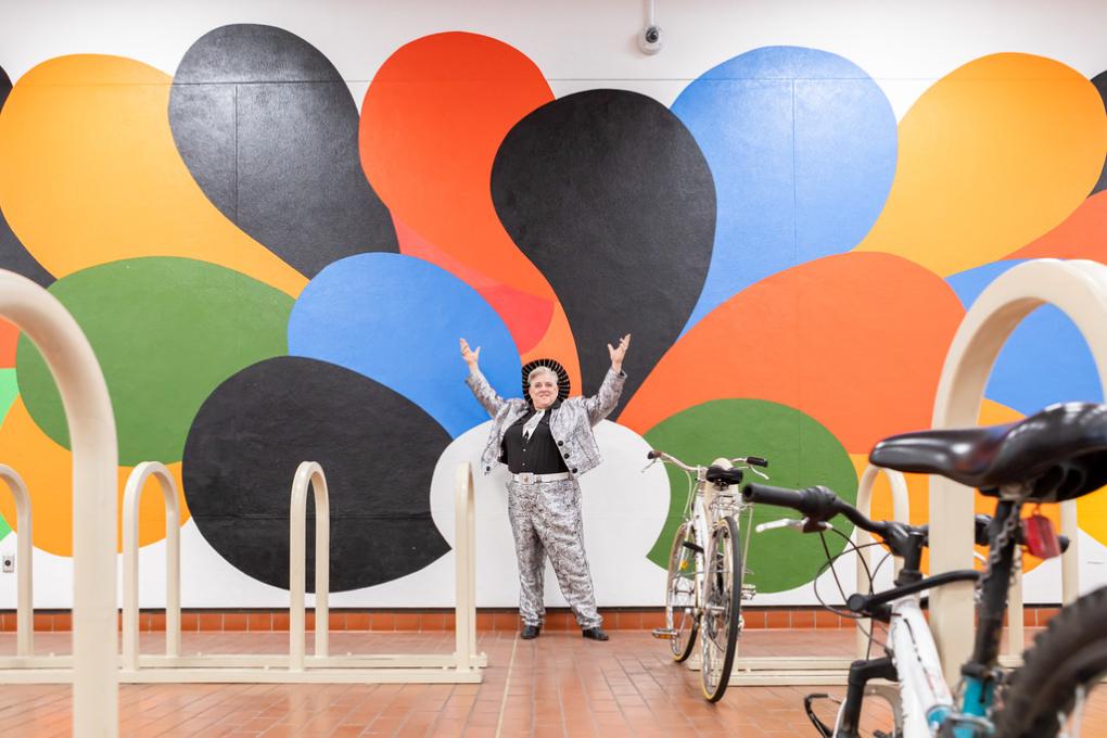 Elvis Herselvis with her hands thrown up in front of a mural with bursts of colors at MacArthur Station