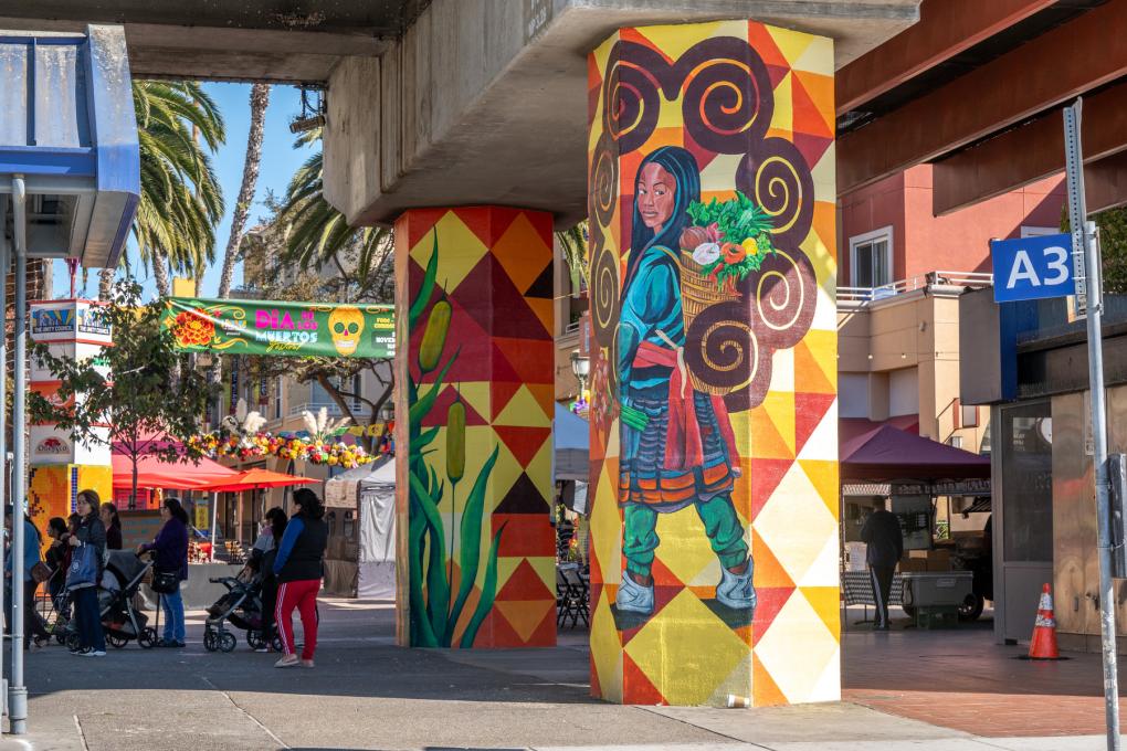 Colorful mural of a person carrying a basket of flowers, painted on a column under a bridge, with a bustling street market visible in the background.