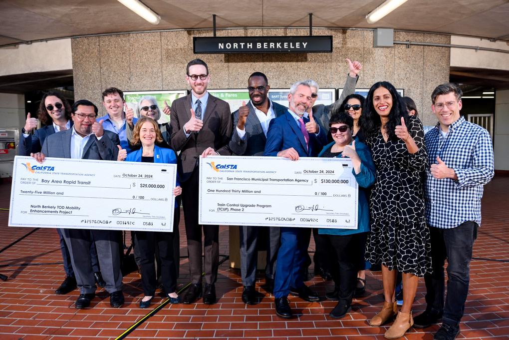 Group of individuals posing joyfully in front of North Berkeley BART station with two oversized ceremonial checks from BART and SFMTA, celebrating a grant for the Bay Area Regional Rail Program.